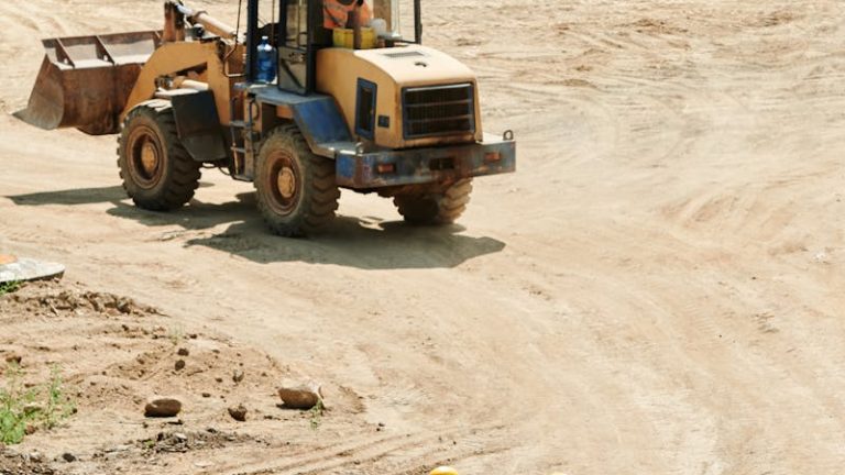 Construction workers in safety gear walking at a busy site with a backhoe in operation.